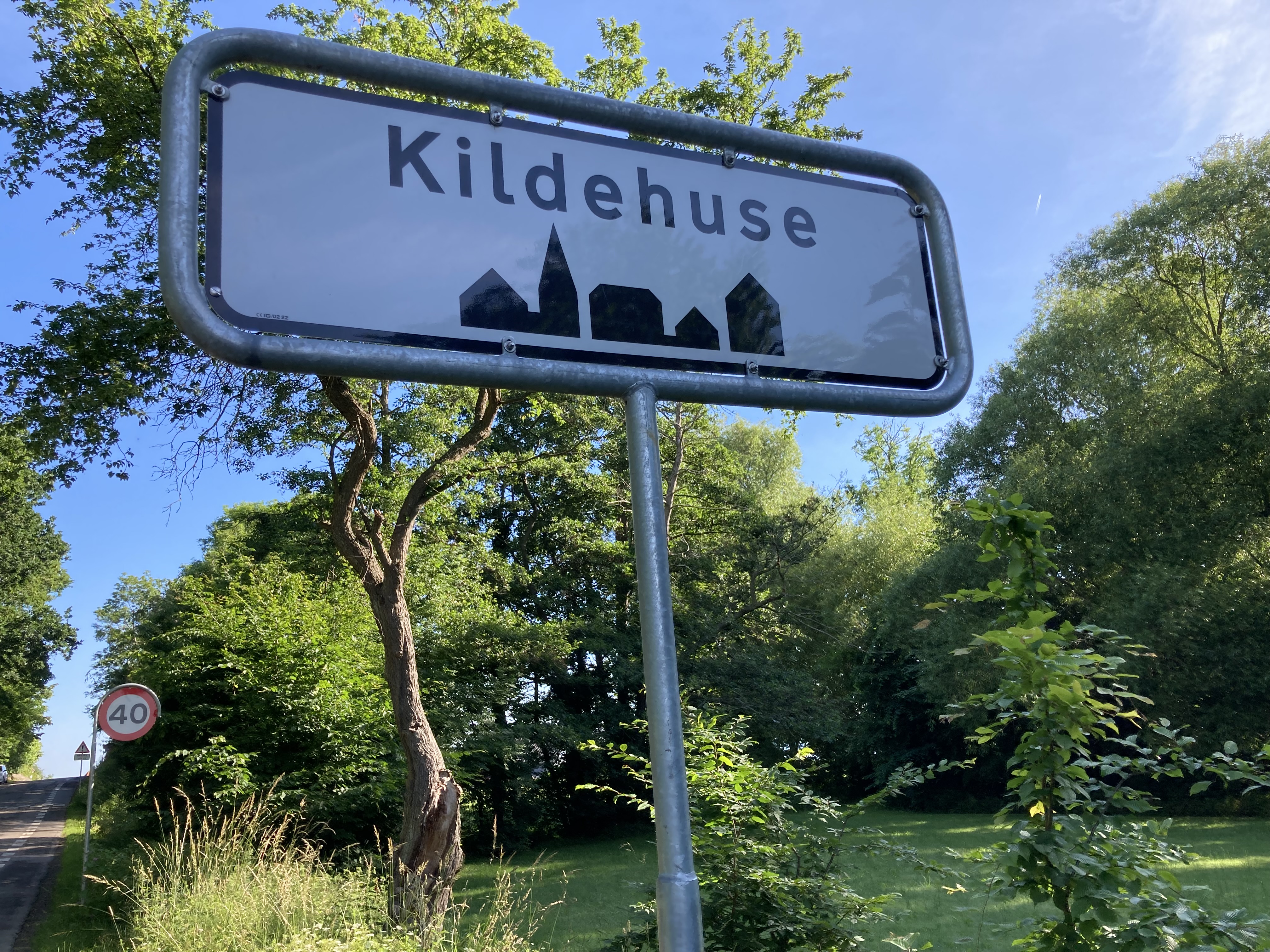 Vejskilt / byzonetavle med teksten "Kildehuse" med solbeskinnede træer i baggrunden